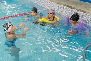 Female instructor guiding diverse children in a indoor swimming pool, practicing various floating and swimming techniques